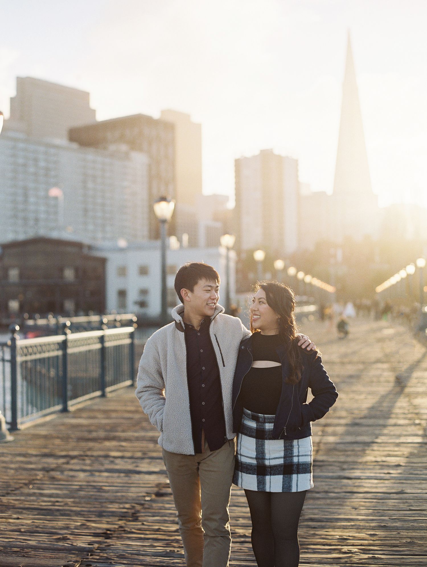 Couple walking together on a waterfront pier at sunset with San Francisco skyline and Transamerica Pyramid in background.