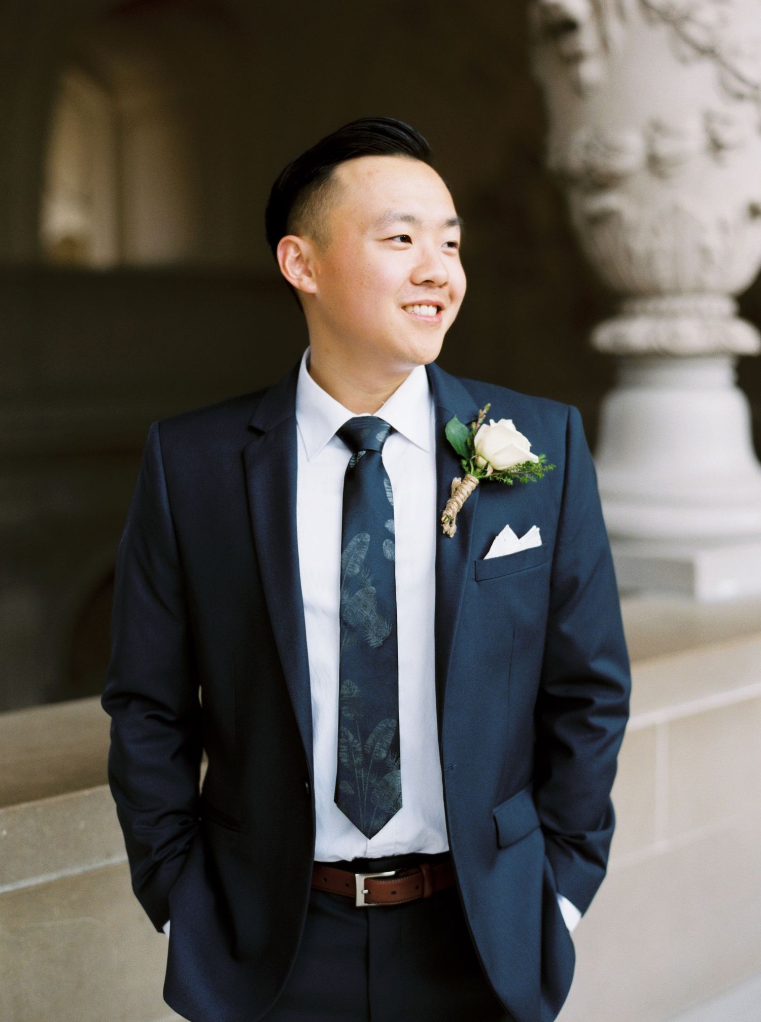 A groom in a navy suit with a white boutonniere smiles while standing in front of architectural columns.