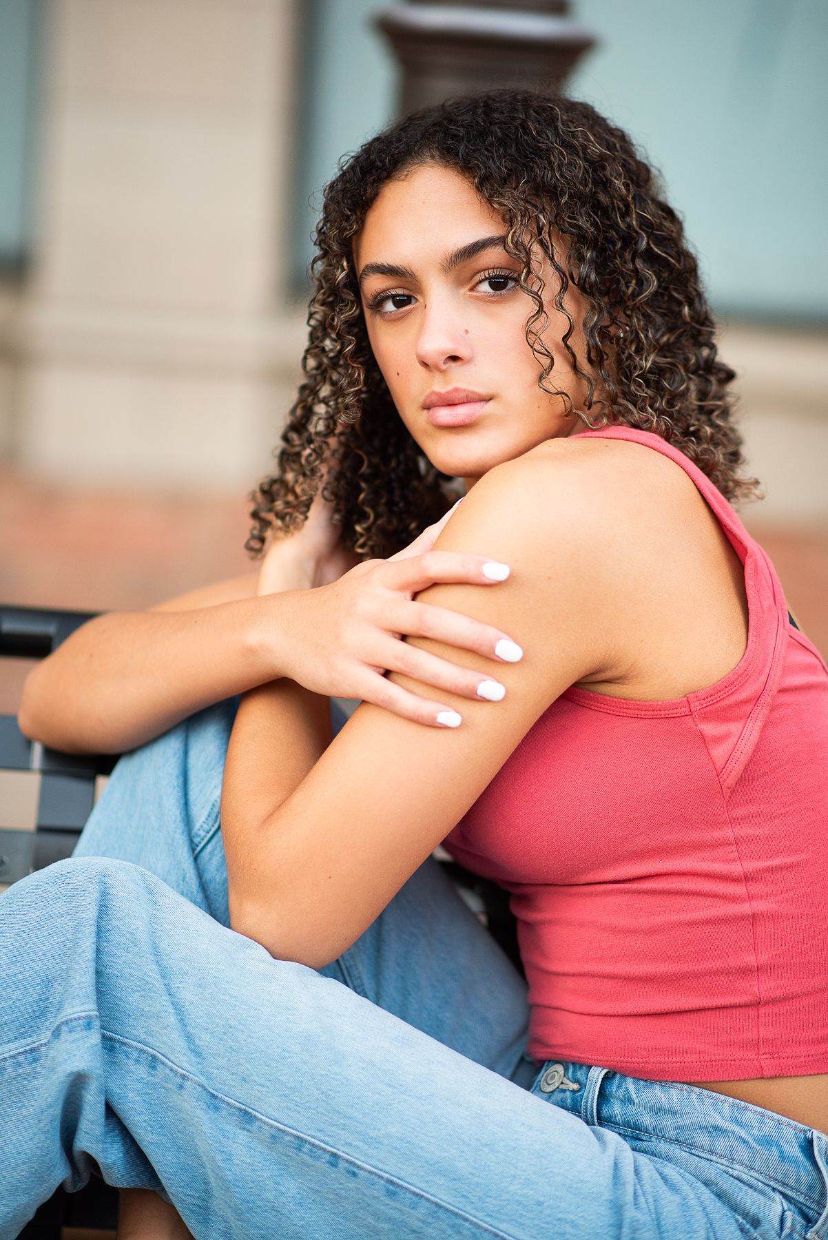 Young woman with curly hair in pink tank top and jeans sitting outdoors, looking at camera with a calm expression.