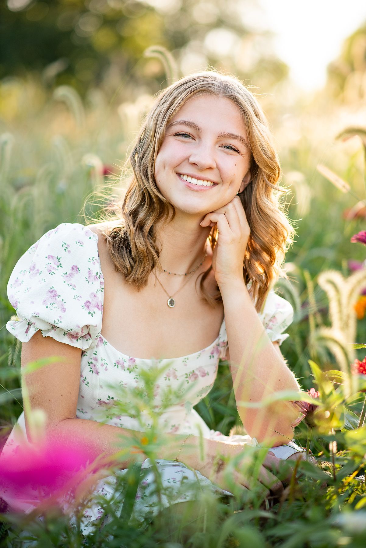 Young woman in floral dress smiling among wildflowers in golden sunlight.