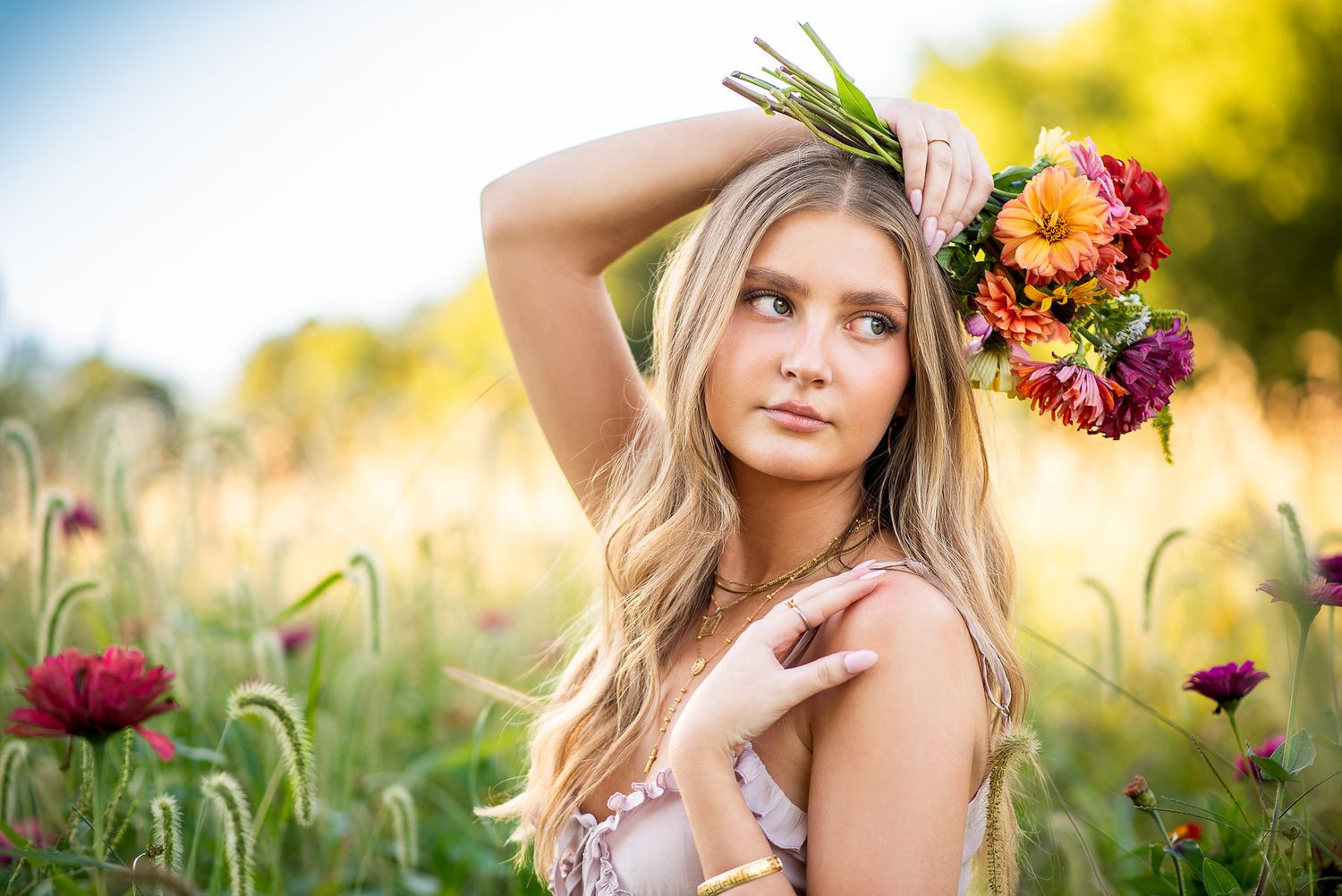 Young blonde woman wearing a colorful floral crown poses gracefully in a sunlit wildflower meadow.