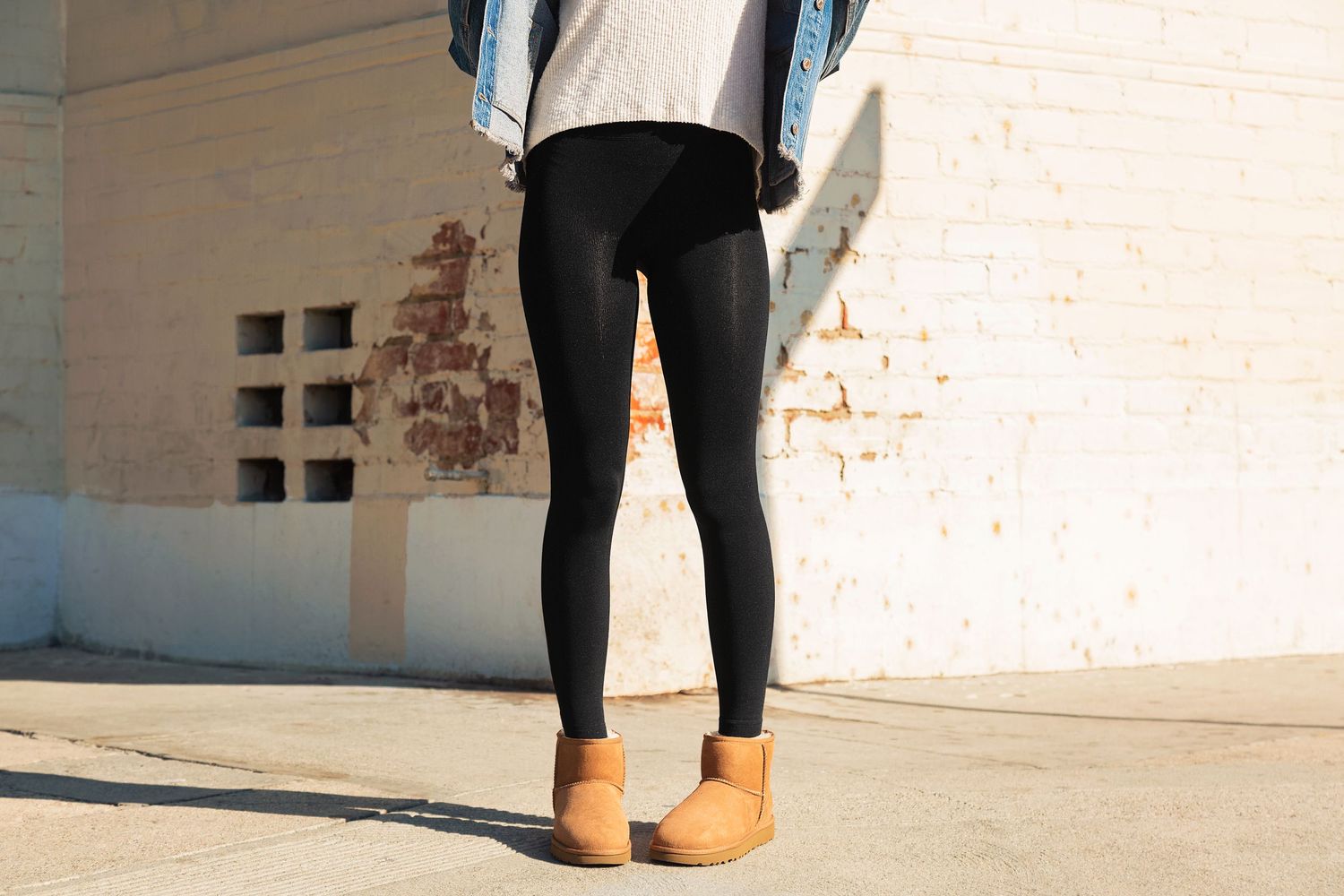 Person wearing black leggings and brown ankle boots stands against a white wall with exposed brick detail.