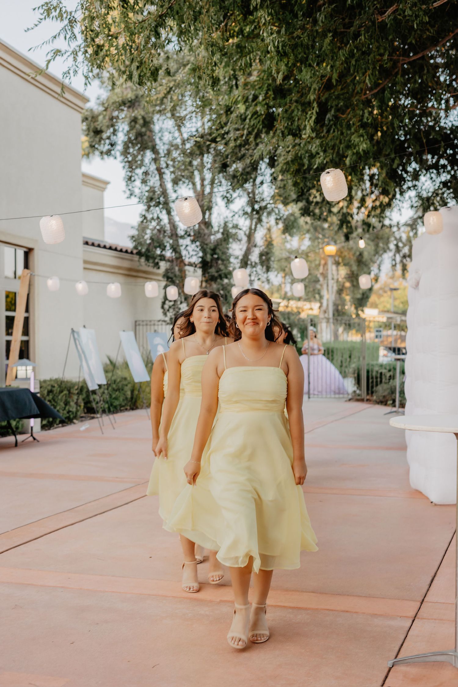 Bridesmaids in pale yellow dresses walk down an outdoor courtyard decorated with hanging lanterns at dusk.