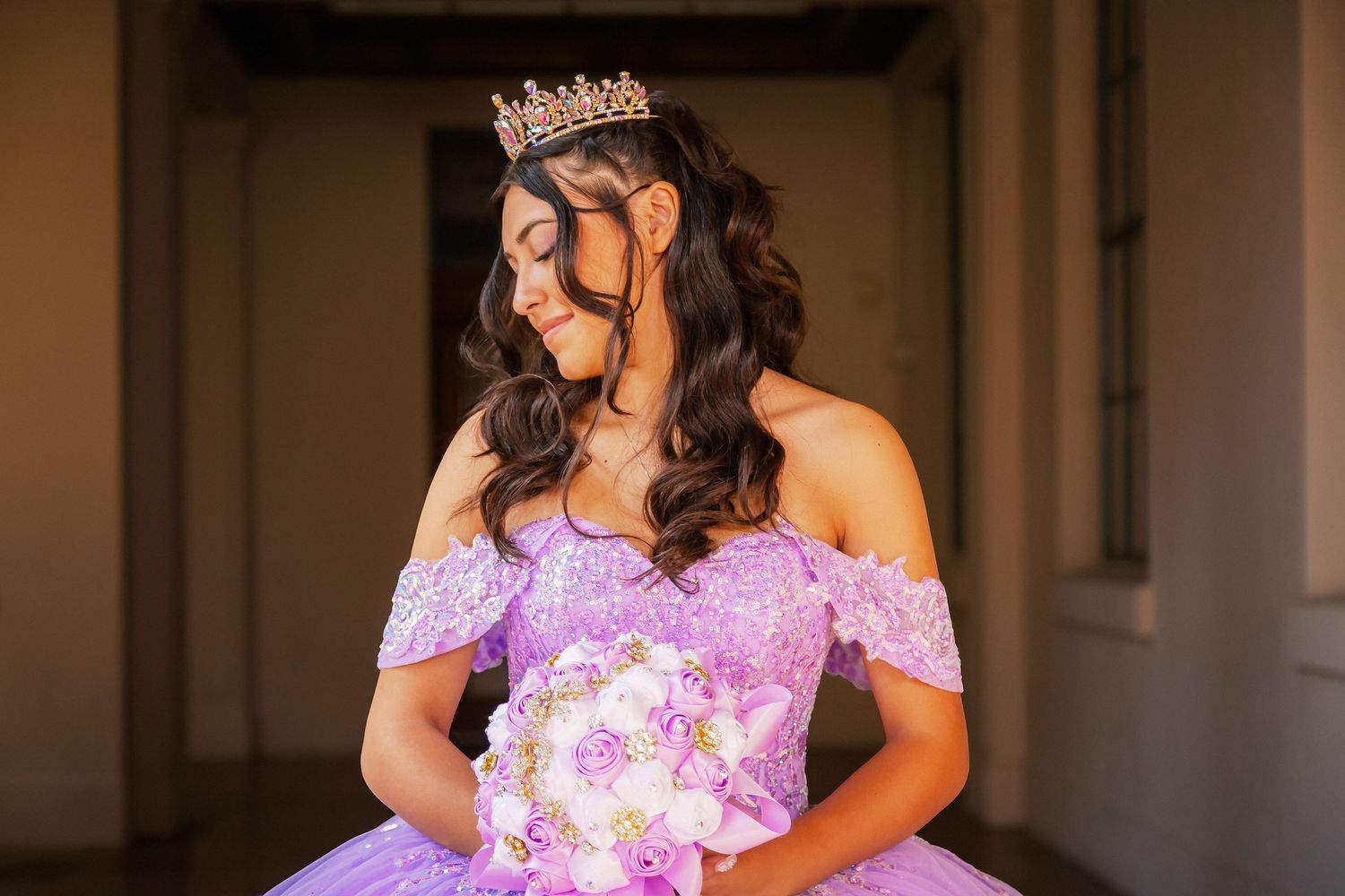 A person wearing a lavender off-shoulder lace gown poses elegantly in an indoor setting.