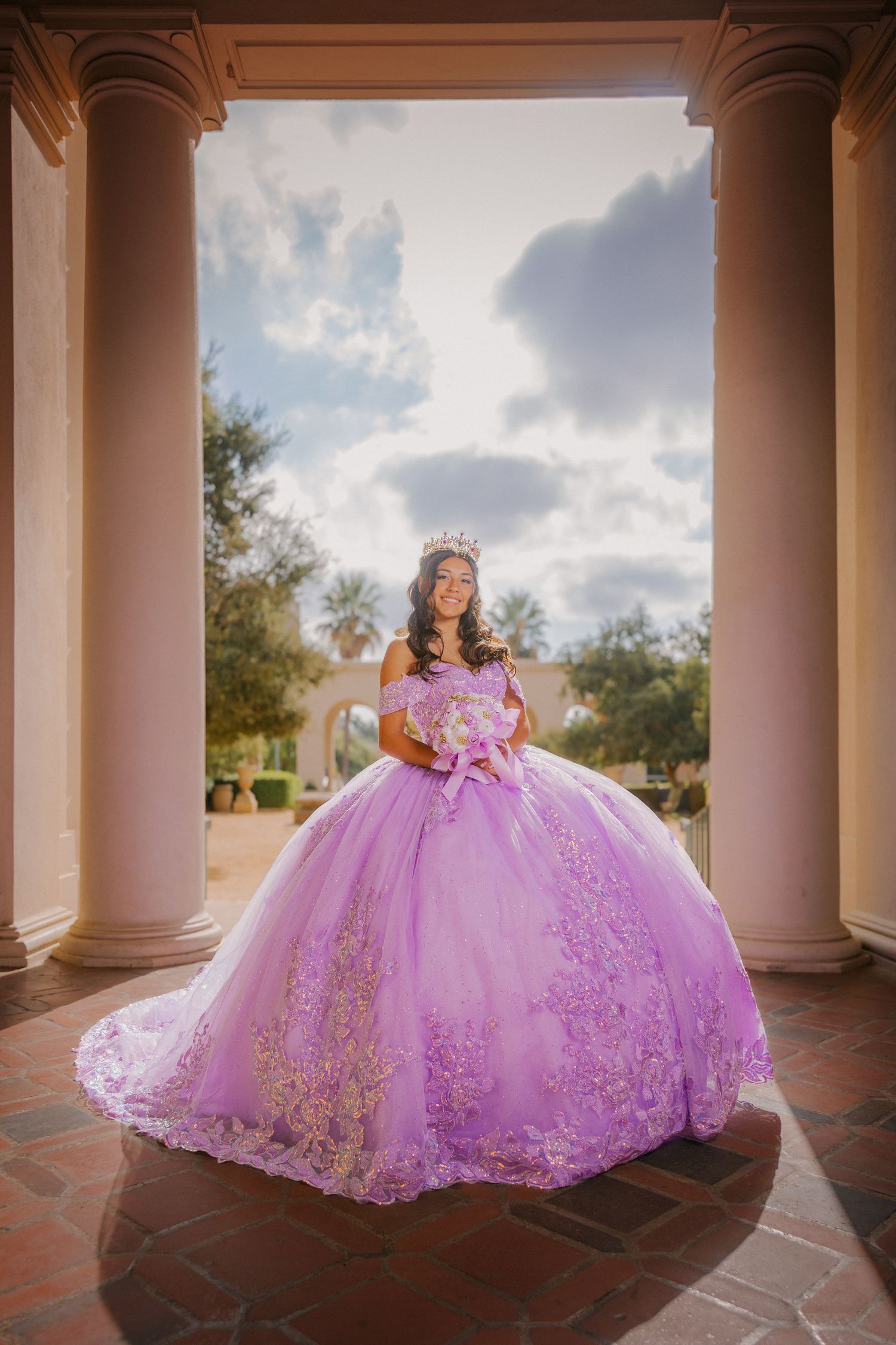 A person wearing a voluminous pink ballgown with gold embellishments poses elegantly between classical columns.