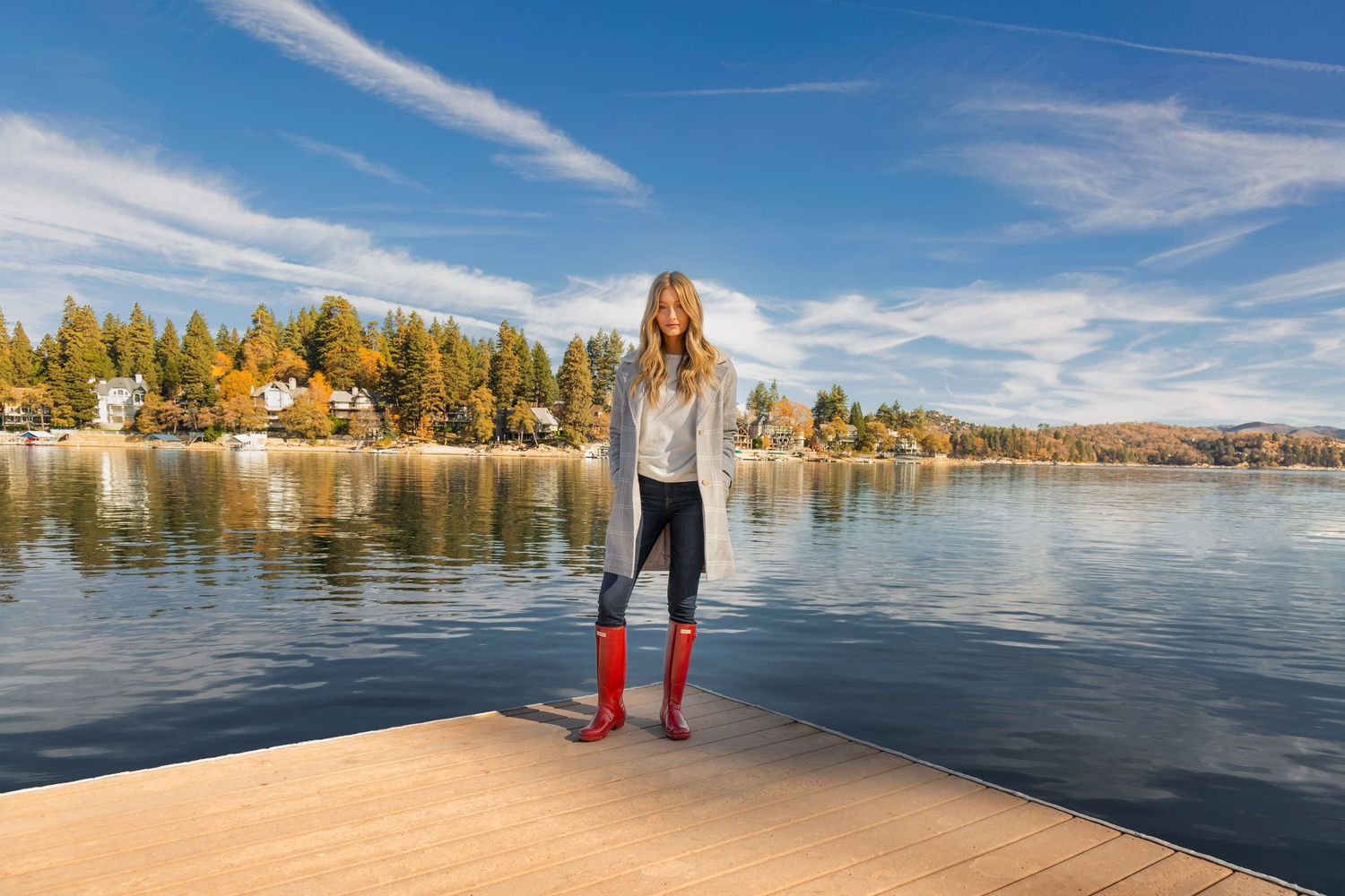 Someone wearing red rain boots stands on a wooden dock overlooking a serene lake with autumn trees and houses along the shore.