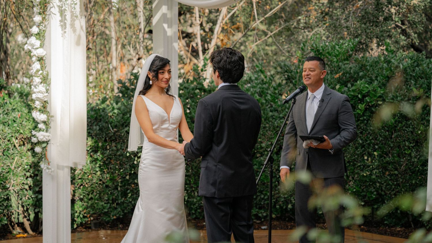 A couple exchanges vows during an outdoor garden wedding ceremony under white columns surrounded by lush greenery.