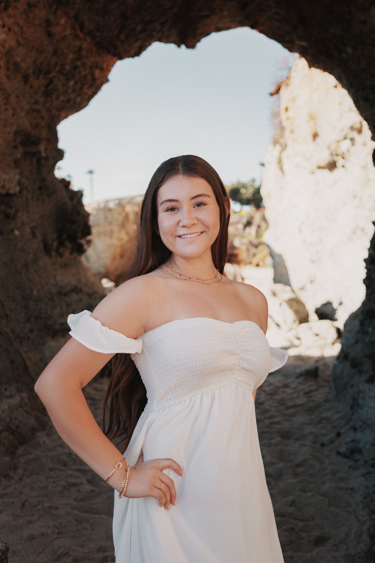 A person in a flowing white dress poses outdoors against a rocky backdrop during golden hour lighting.