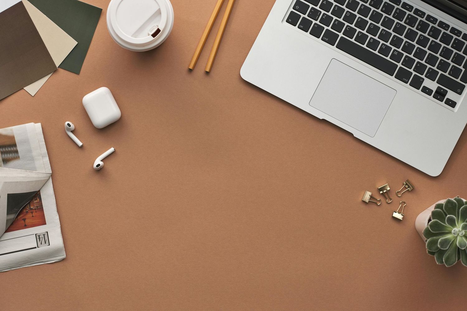 Modern workspace flatlay with laptop, wireless earbuds, coffee cup and office supplies on brown desk surface.