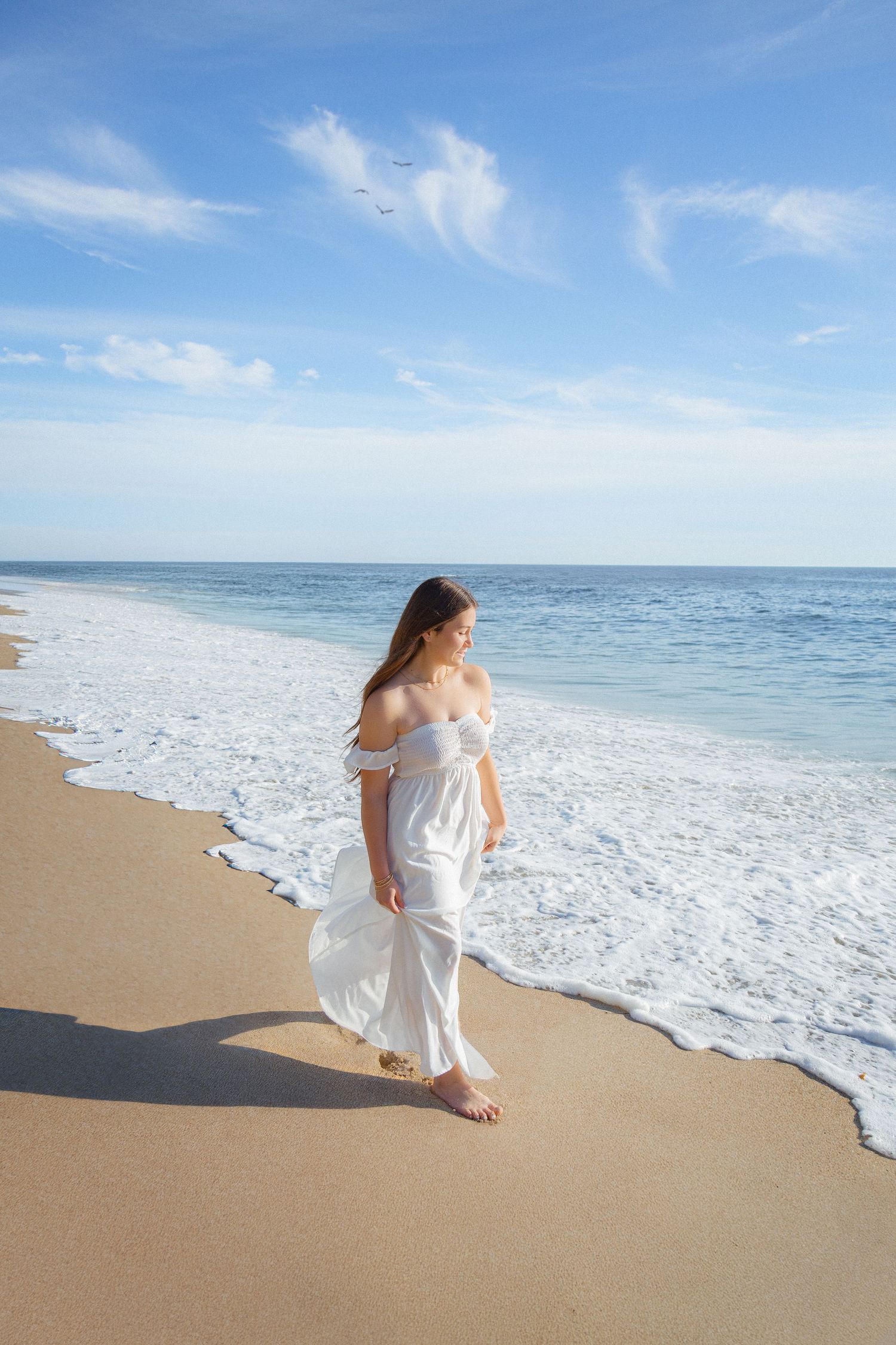 A person in a flowing white dress walks barefoot along a pristine beach shoreline on a sunny day.