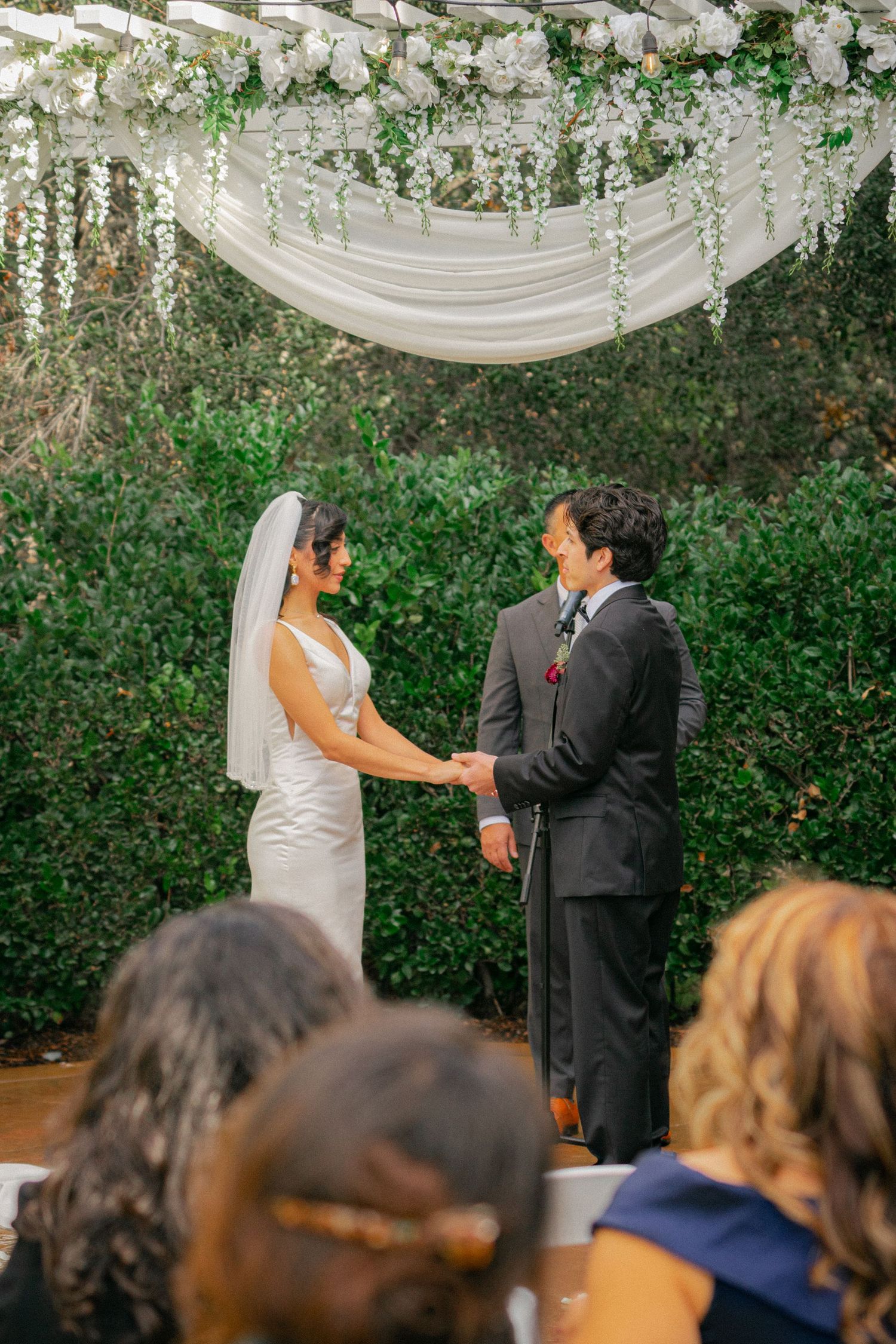 A wedding ceremony takes place under elegant white draping and greenery at an intimate outdoor venue.