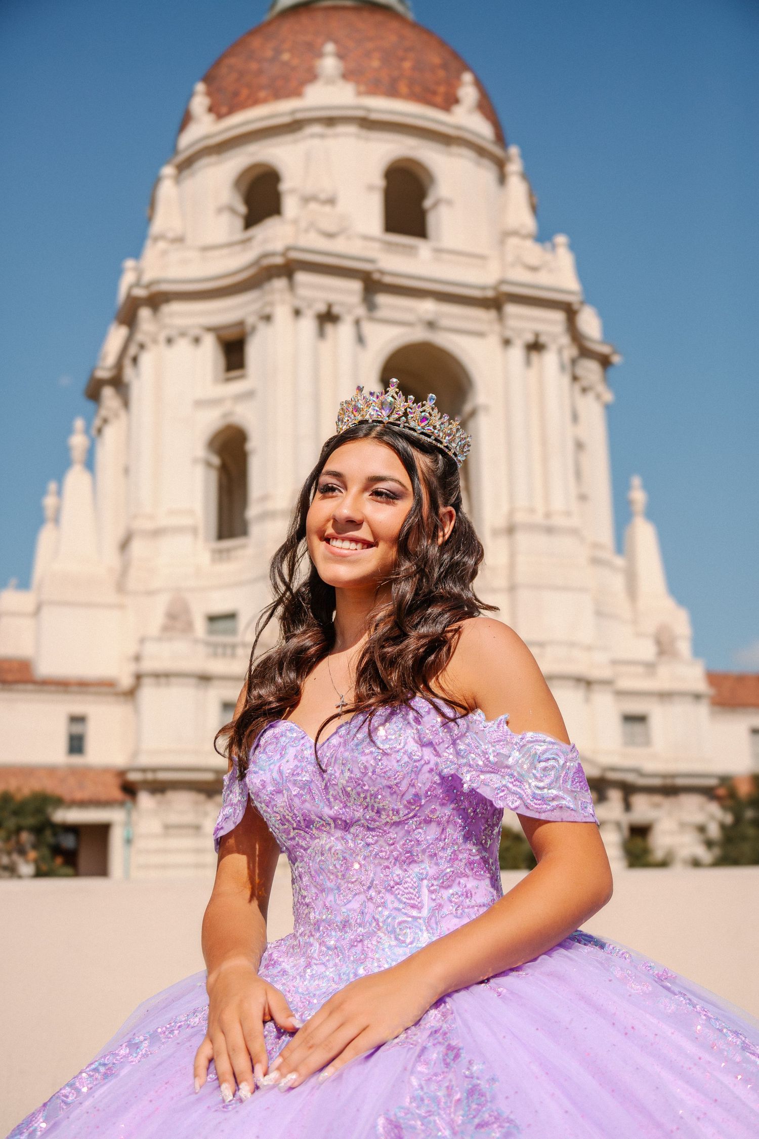 A person in a lavender quinceañera dress poses in front of a grand white cathedral building on a sunny day.