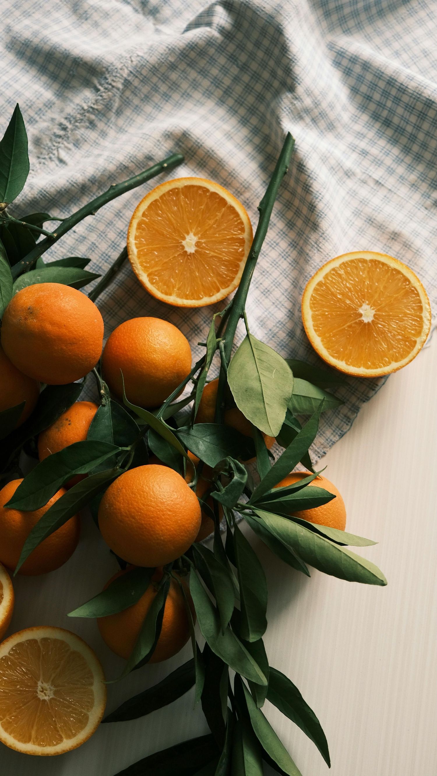Fresh oranges and orange halves arranged with green leaves on white textured fabric in natural lighting.