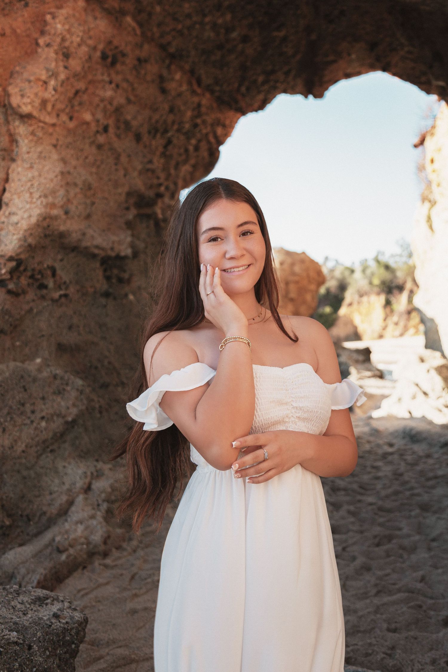A person in a white off-shoulder dress poses against a rocky cave opening with natural lighting.