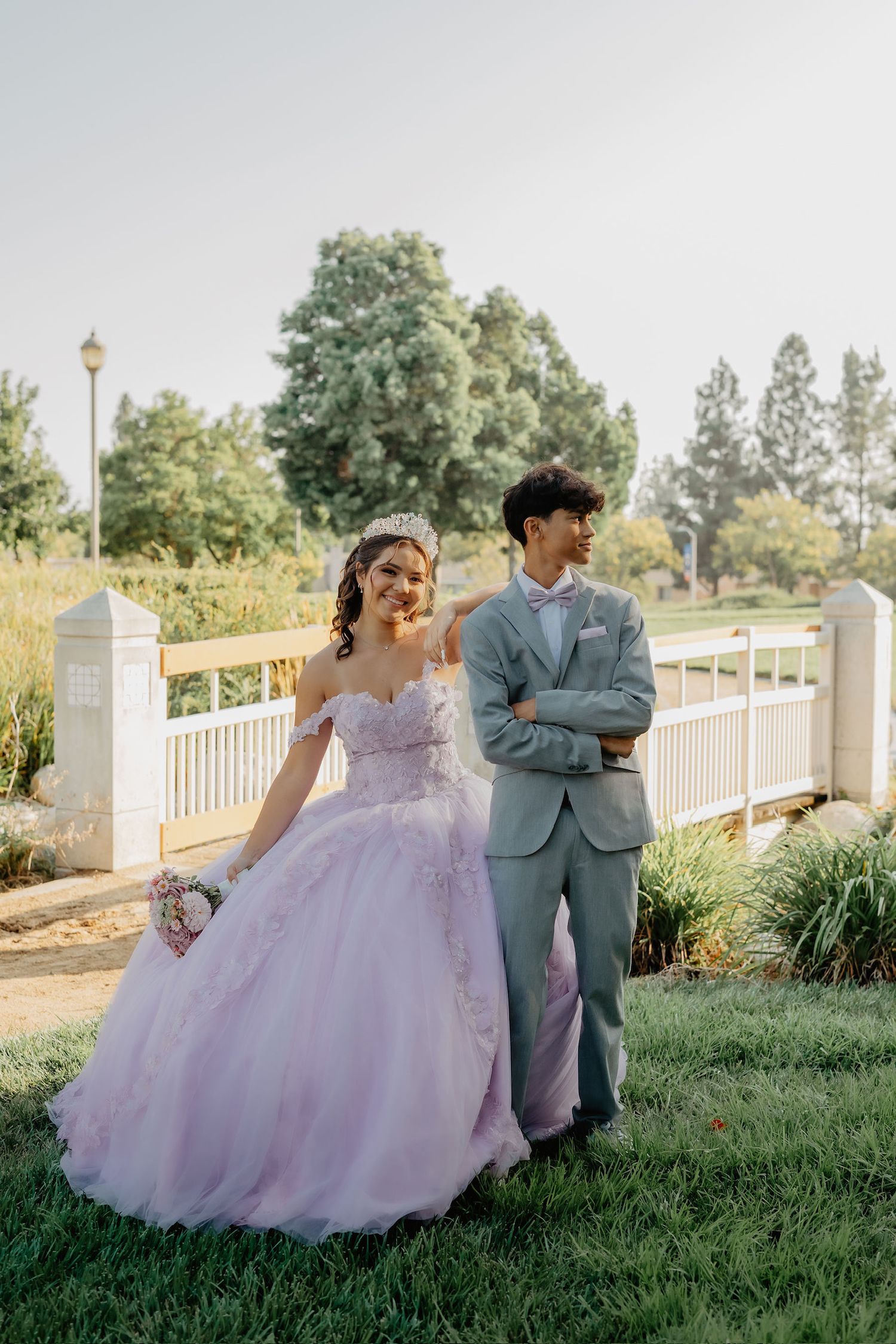 A couple in formal attire poses playfully on a grassy hill with white fencing and trees in the background.