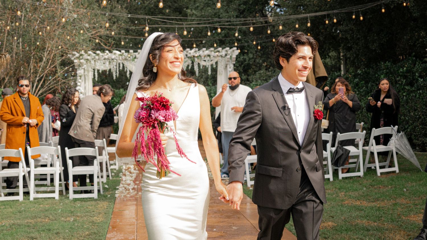 A couple walks down an outdoor wedding aisle with string lights overhead and white chairs for guests.