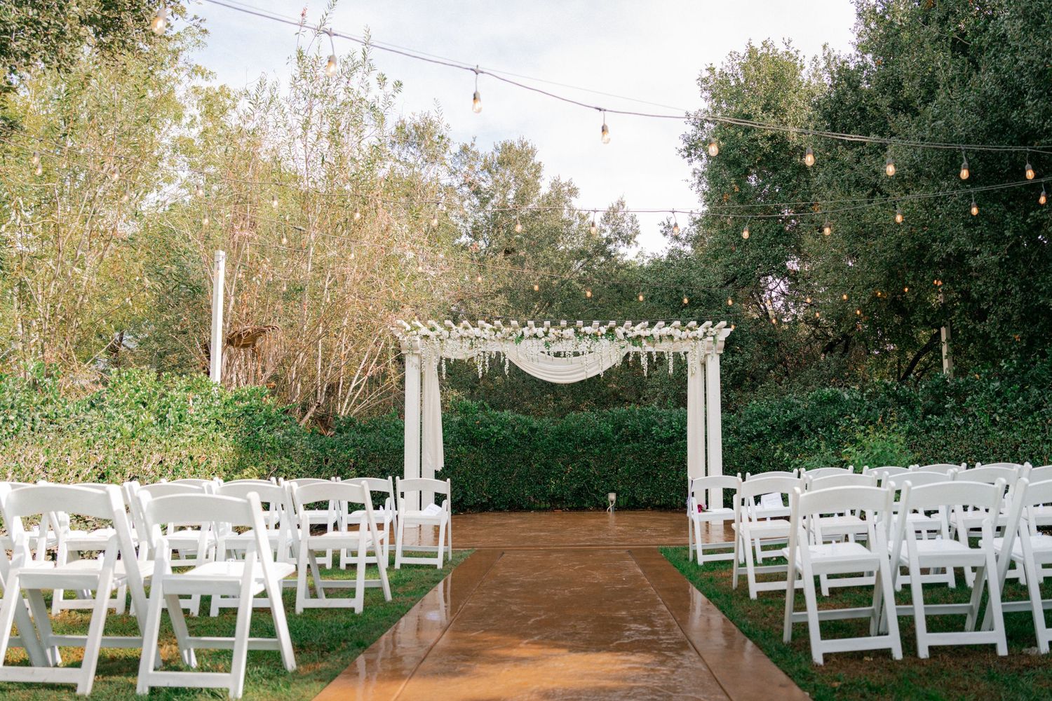 White wooden chairs arranged in rows for an outdoor garden wedding ceremony under string lights and natural greenery.