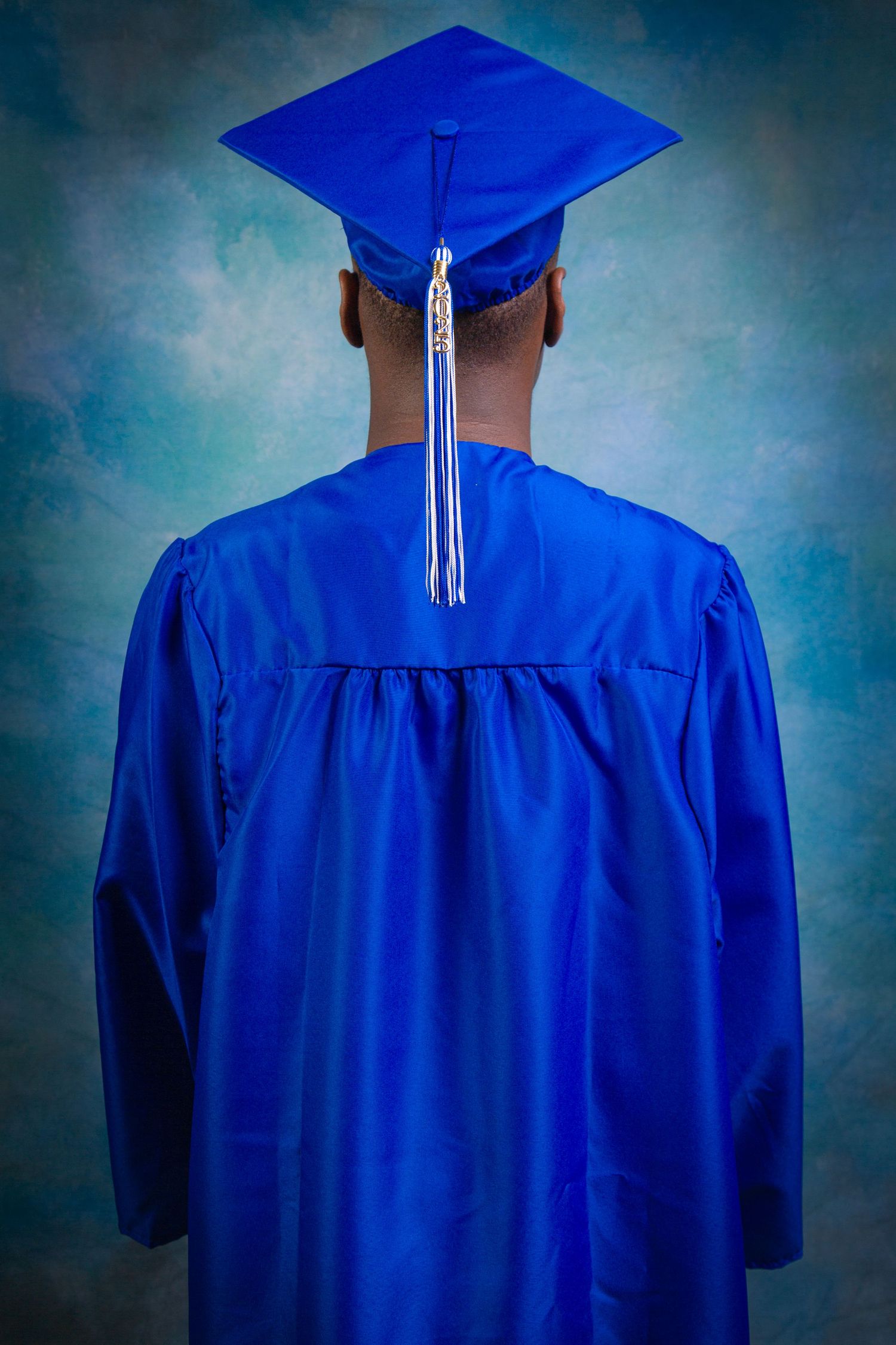 Back view of graduate wearing bright blue cap and gown with white tassel against blue backdrop.