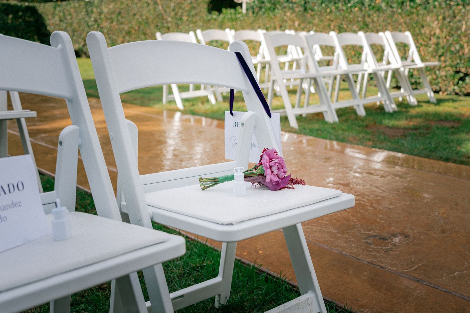 White folding chairs set up for an outdoor wedding ceremony with a pink flower bouquet resting on one chair seat.
