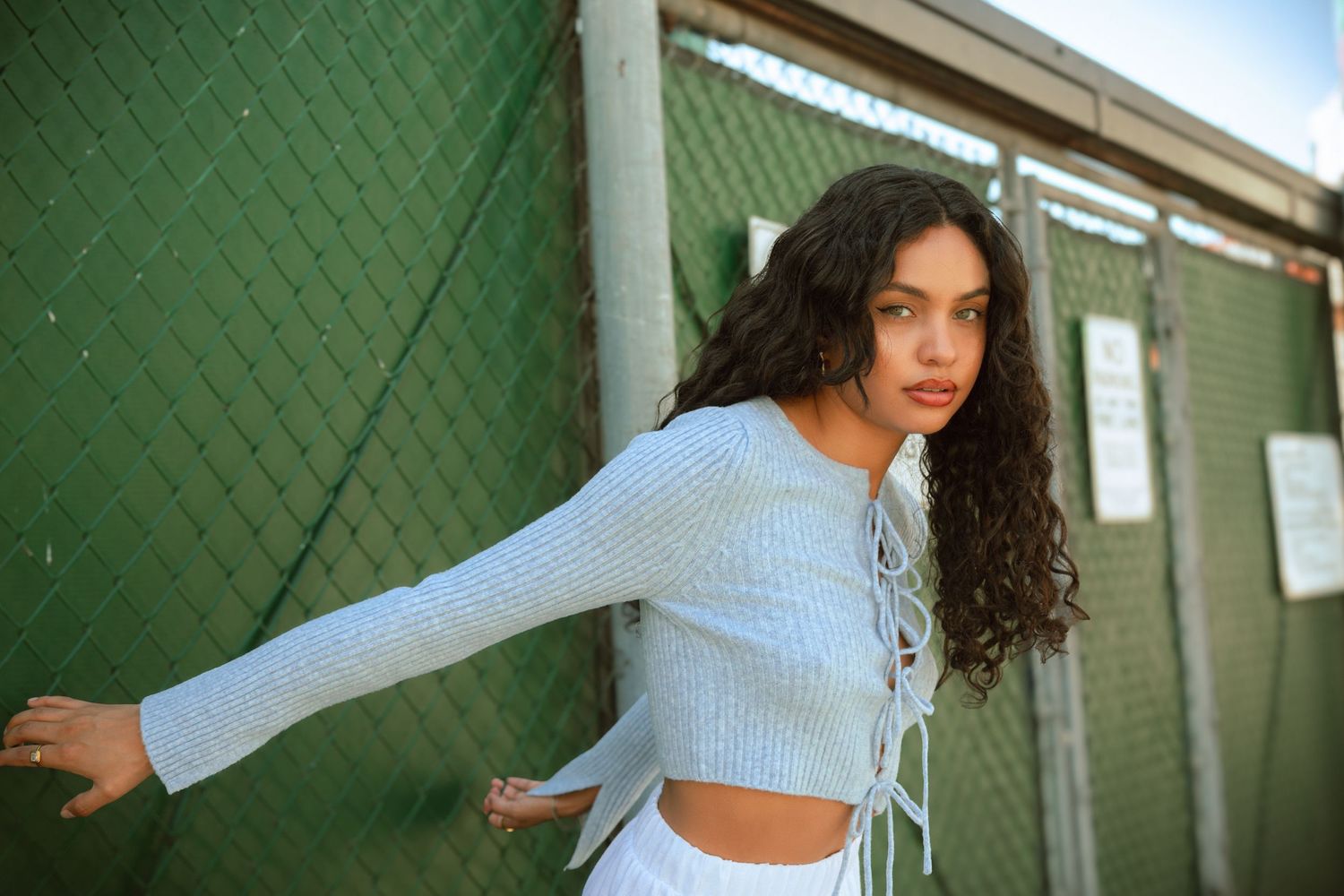 Woman in light blue crop top poses playfully against green wall in urban setting.