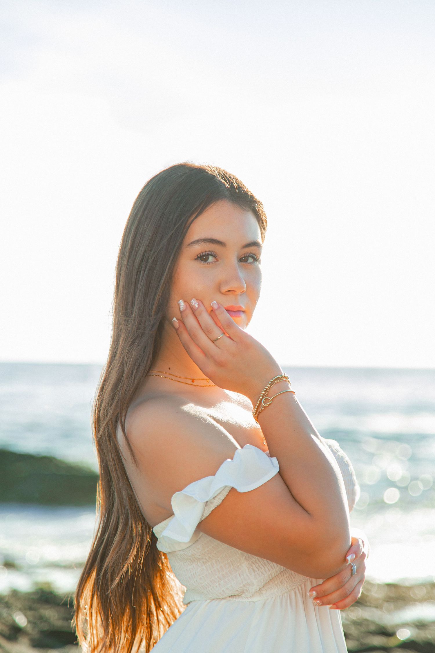 A woman in a white dress poses at sunset on a beach with golden sunlight and ocean waves in the background.