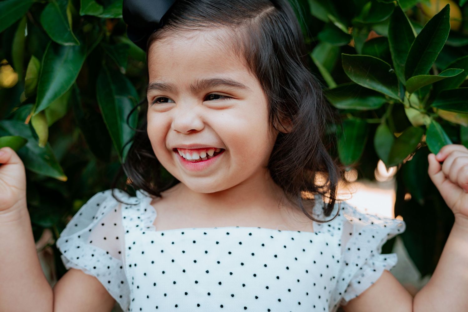 Child in polka dot dress poses joyfully against green foliage background.