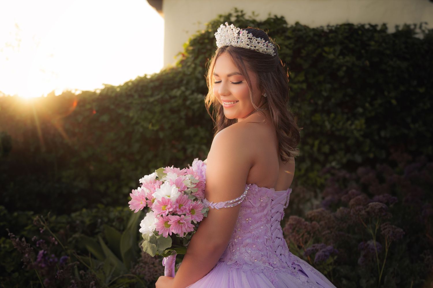 A bride in a lavender gown holds a pink and white bouquet against a sunset backdrop.