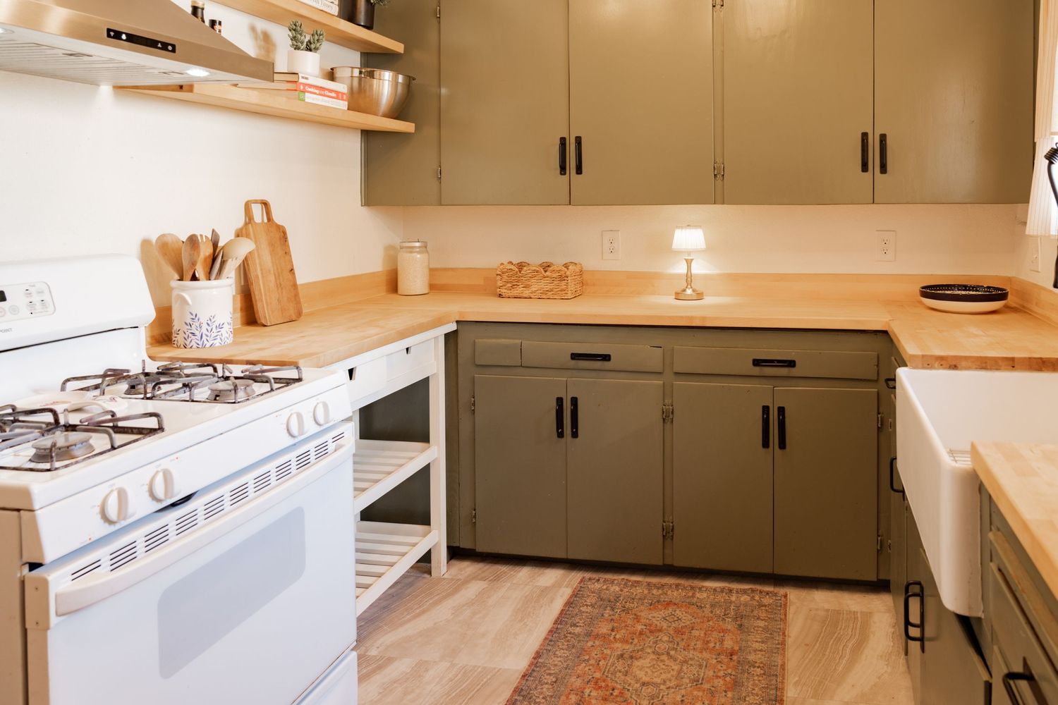 Modern kitchen with olive green cabinets, wood shelving, and patterned area rug on light hardwood floors.
