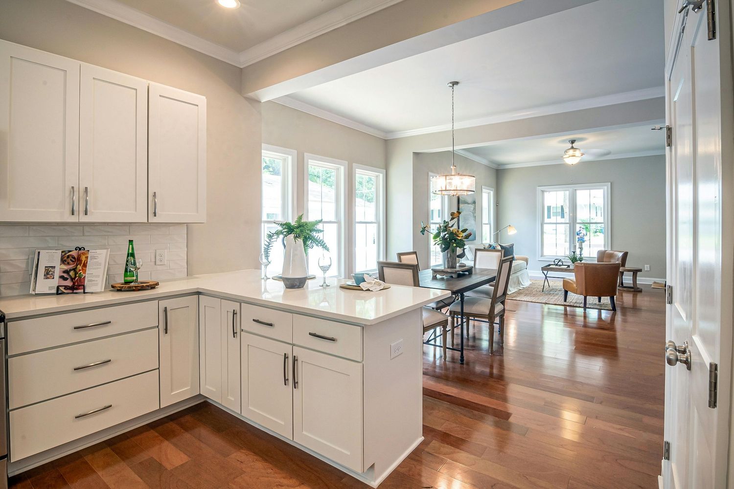 Modern white kitchen with hardwood floors opens to bright dining area featuring pendant lighting and coastal decor.