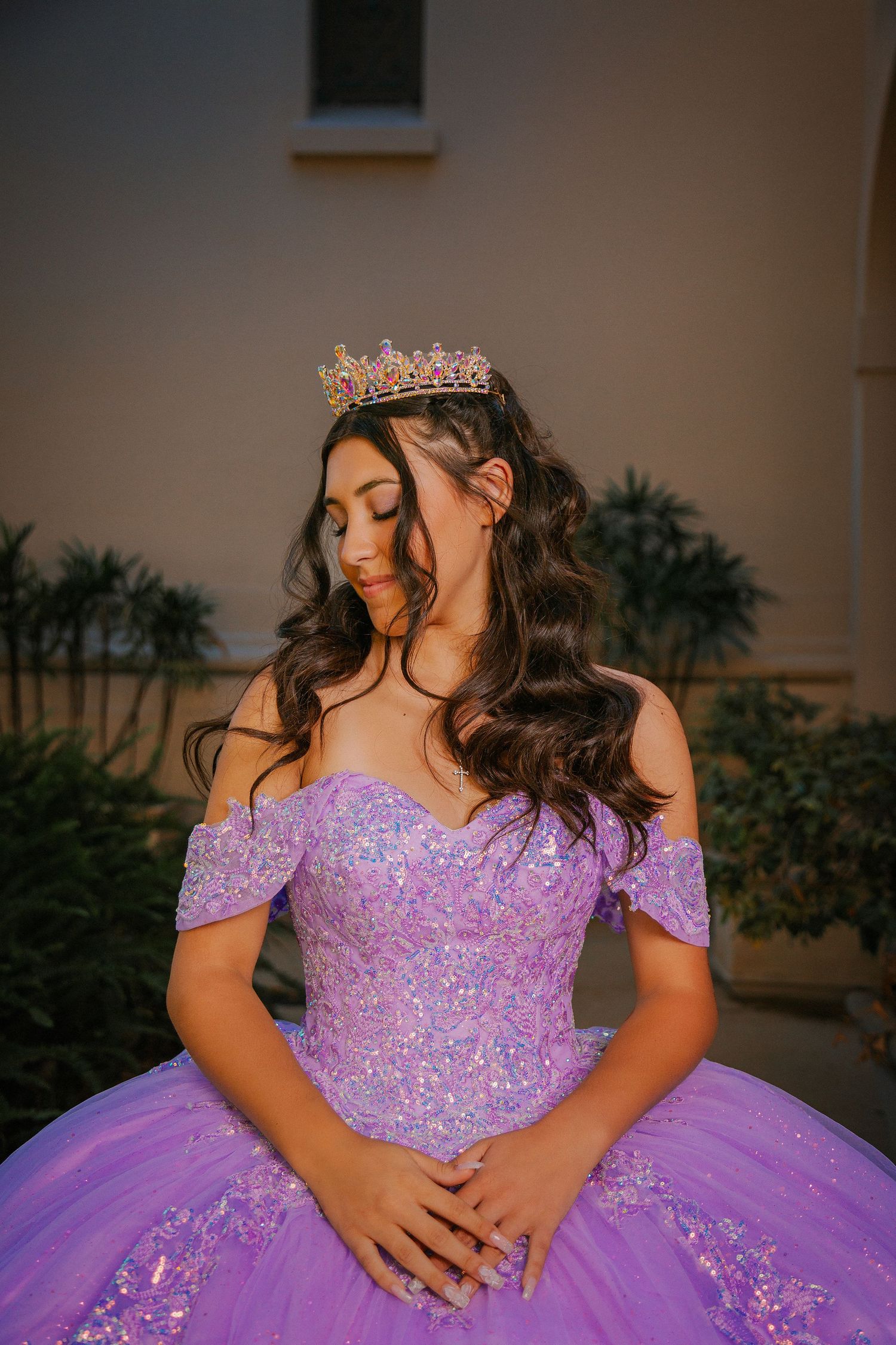 A person in a sparkly purple quinceanera ball gown with off-shoulder sleeves and a tiara poses for photos.