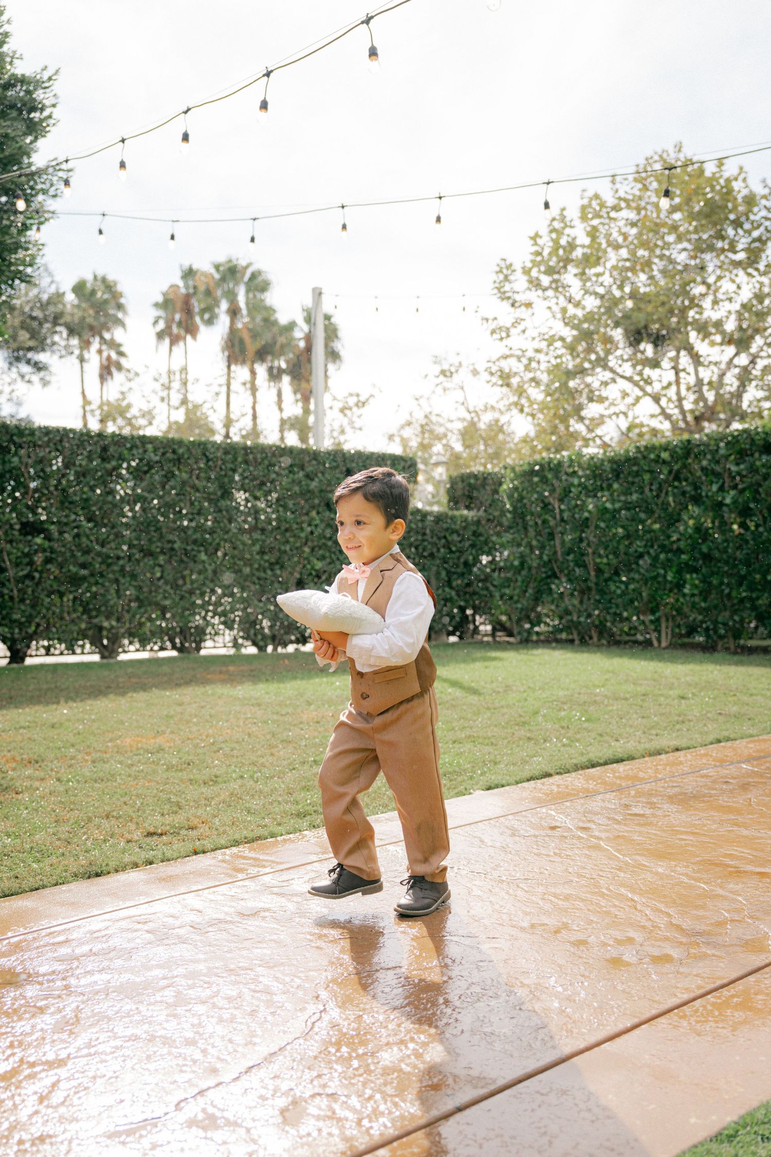A small child in brown overalls splashes and plays on a wet patio under string lights during an outdoor celebration.