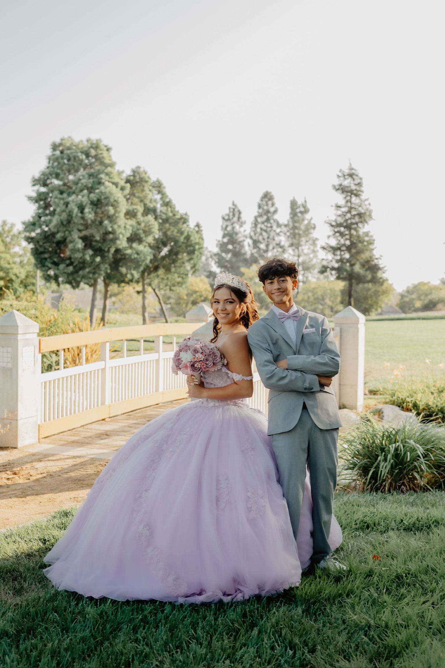 A couple in formal attire poses together on a grassy hill with a white fence and trees in the background.