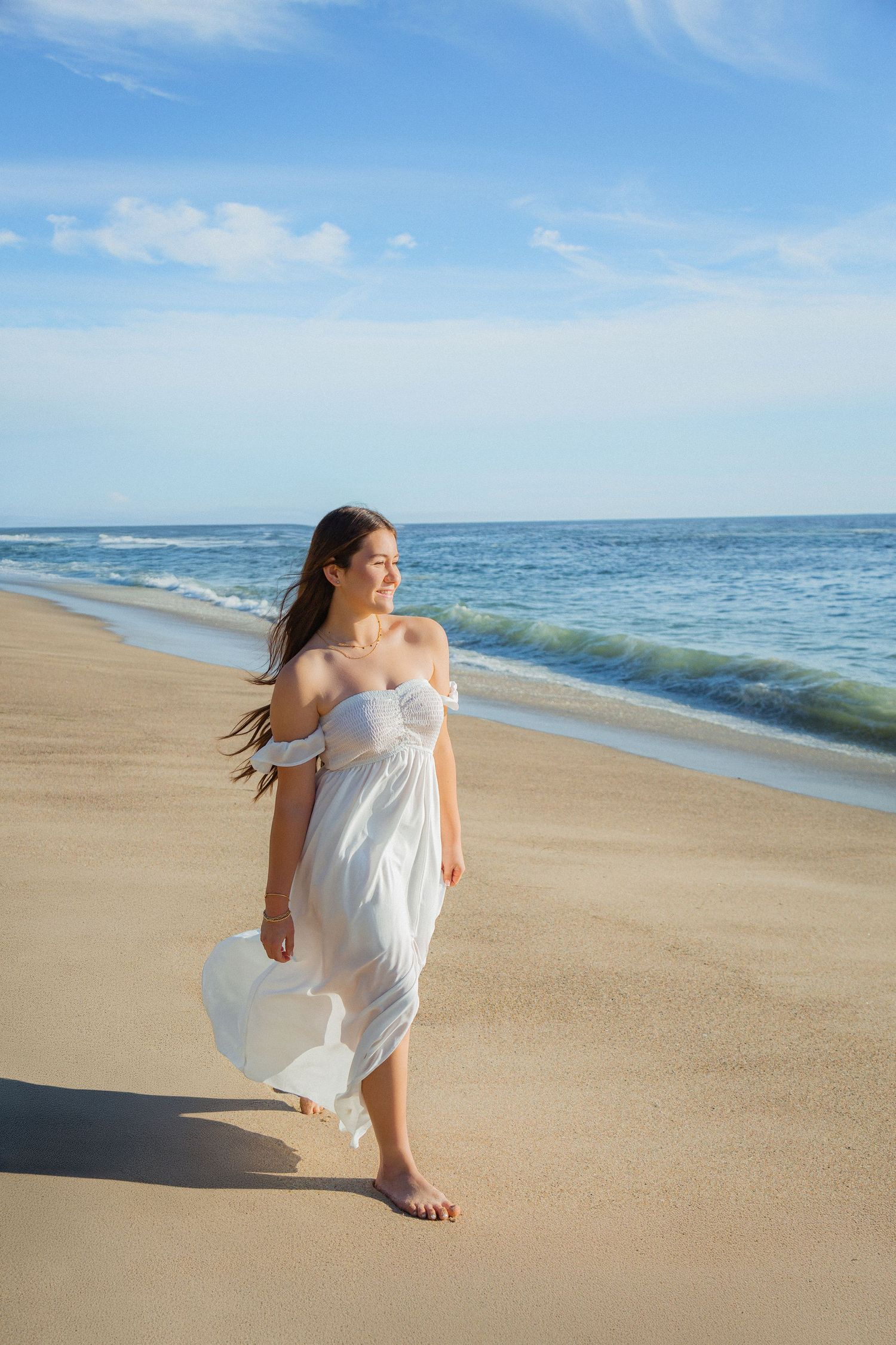 A graceful figure in a flowing white dress dances along a pristine beach shoreline on a sunny day.