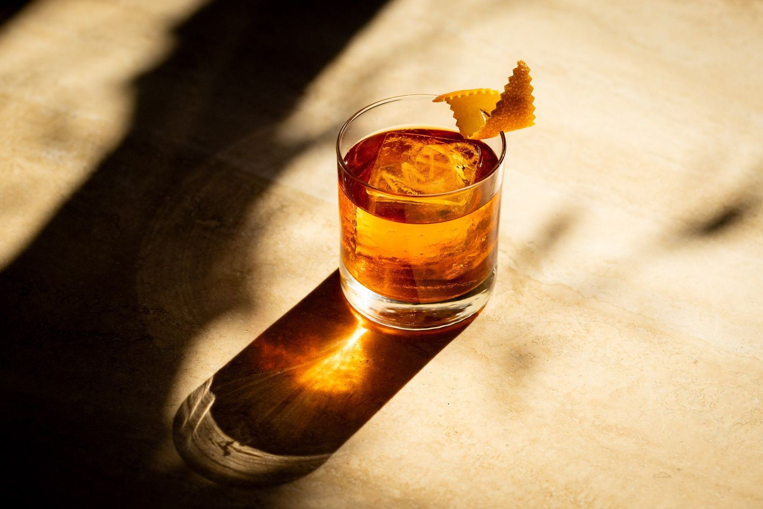 A golden-hued cocktail in a rocks glass garnished with an orange peel sits in dramatic lighting casting a long shadow.