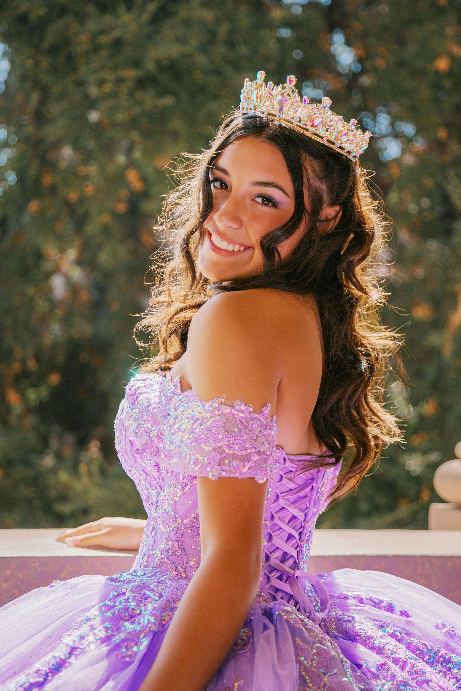 A young woman in a lavender quinceañera dress poses outdoors with sunlit trees in the background.