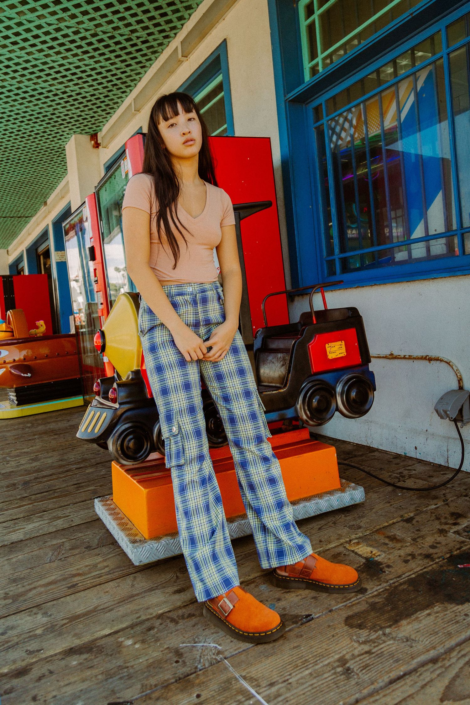 A person in a pink top and plaid pants poses on a boardwalk with red toy trains in the background.