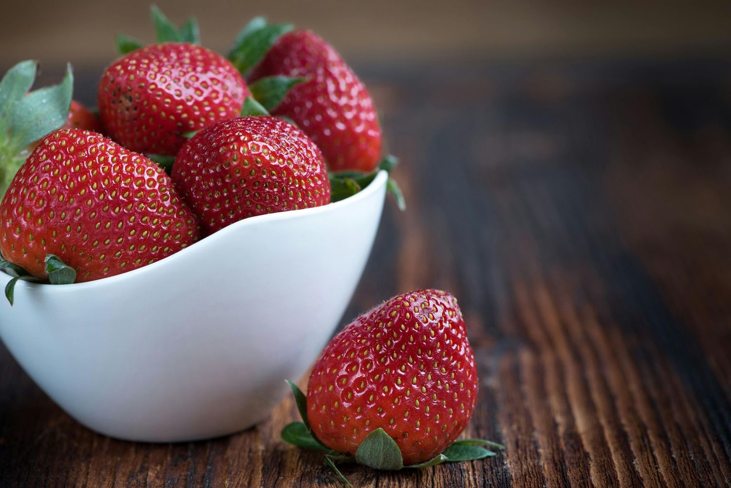 Fresh ripe strawberries arranged in white ceramic bowls on a rustic wooden table surface.
