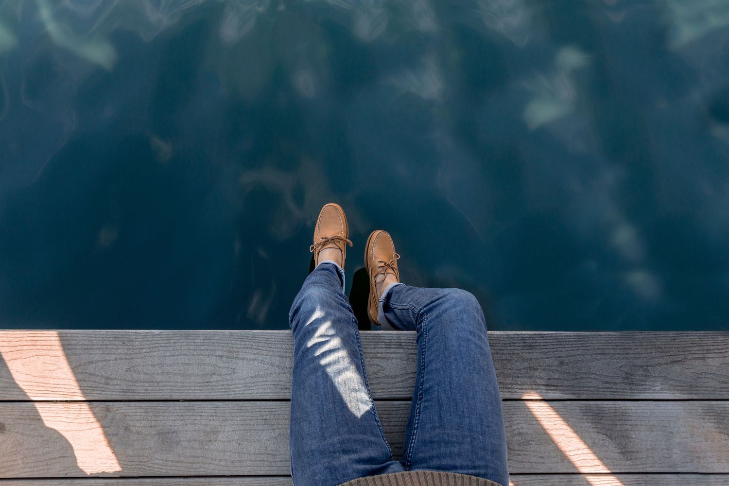 Person wearing blue jeans and brown shoes sitting on wooden dock with dark water below.