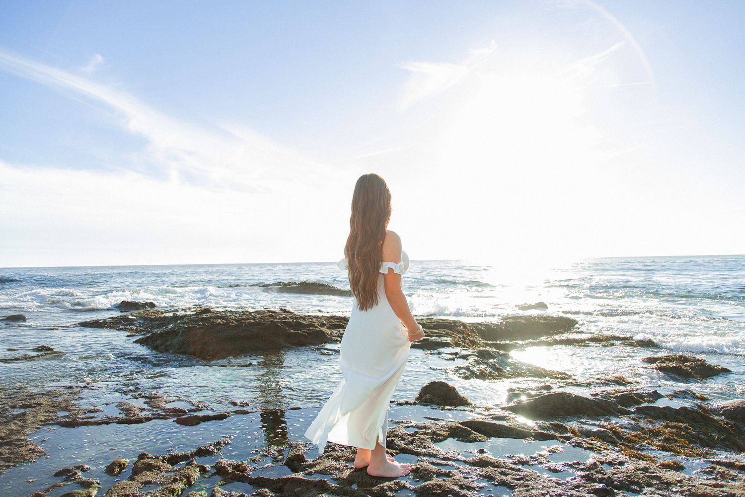 Woman in white dress exploring rocky beach coastline at sunset with ocean waves crashing against shore.