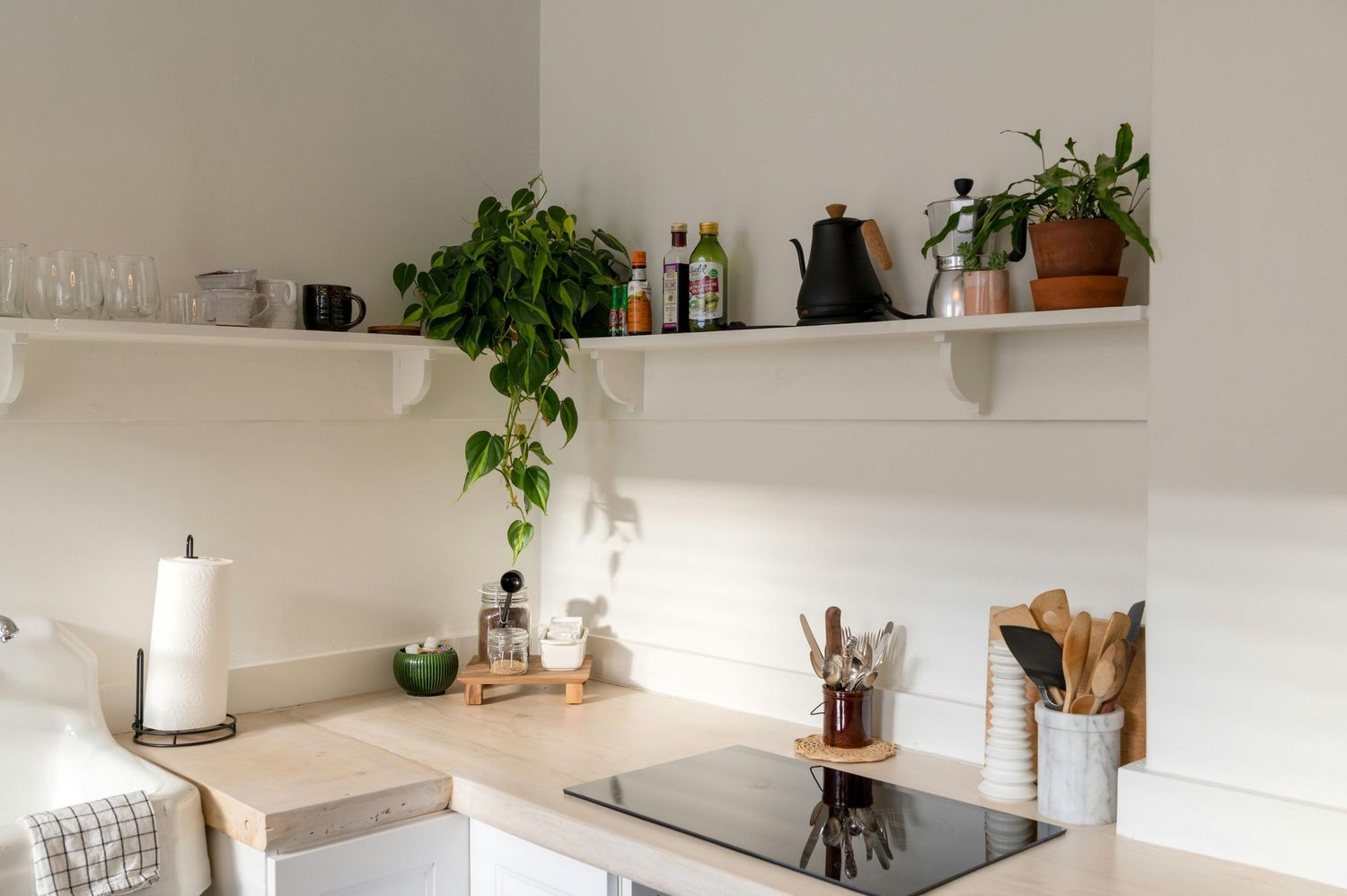 Modern minimalist kitchen with white walls, floating shelves, potted plants, and wooden utensils on a clean countertop.