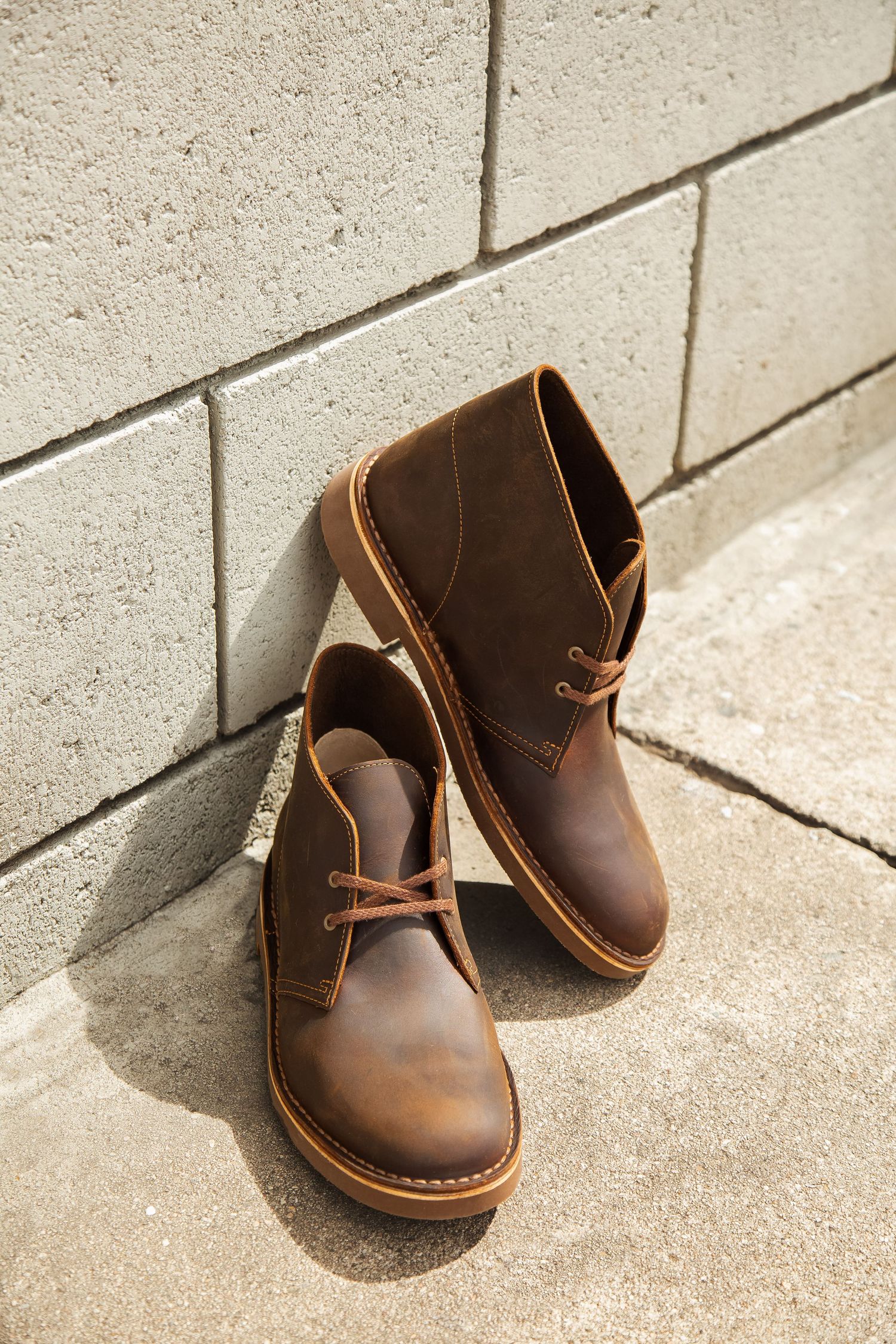 Brown leather chukka boots with contrast stitching displayed on concrete floor against white brick wall.