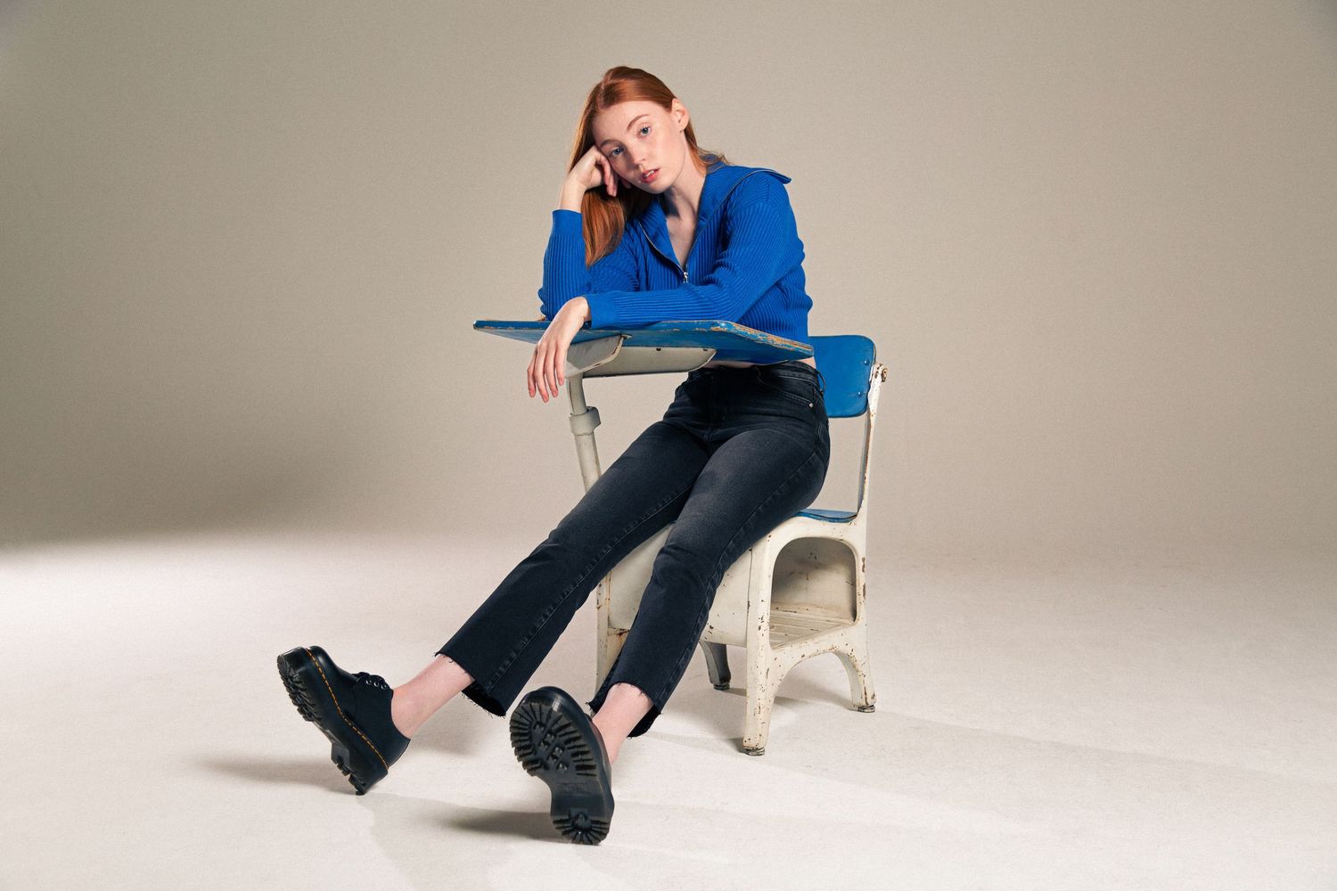 Person in blue shirt and dark pants sits on white chair in studio setting against neutral background.