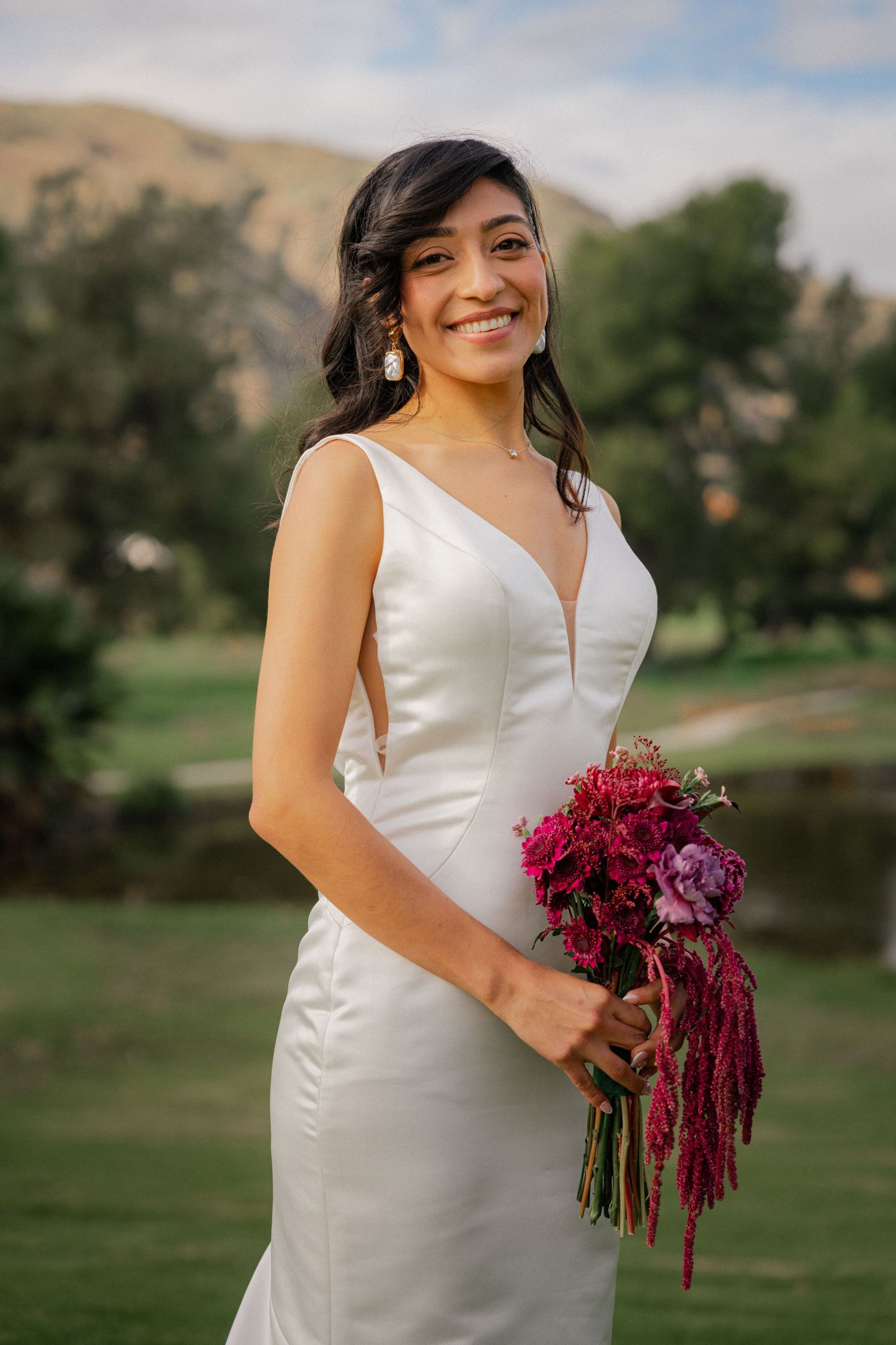 A bride in a sleek white satin dress holds a vibrant burgundy and purple cascading bouquet in an outdoor garden setting.