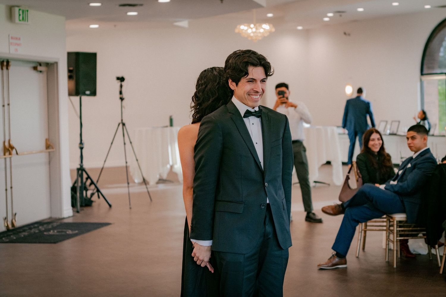 A person in a dark suit and bow tie stands in an elegant event space with guests seated in the background.