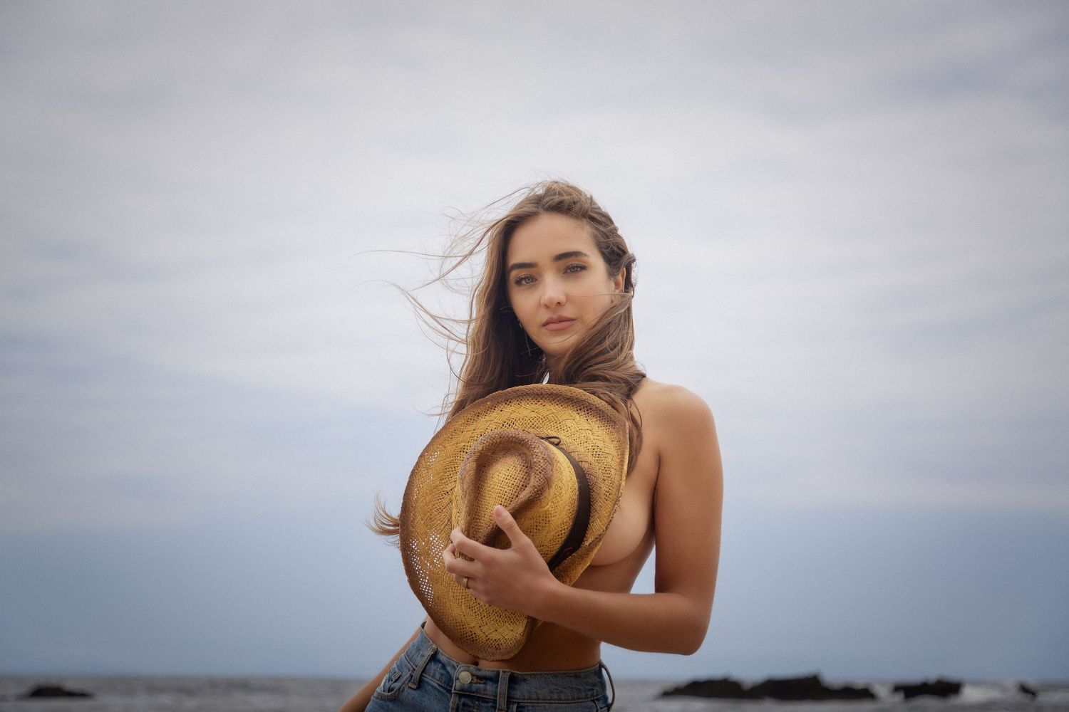 A woman in a straw hat and black swimsuit poses on a beach during golden hour.