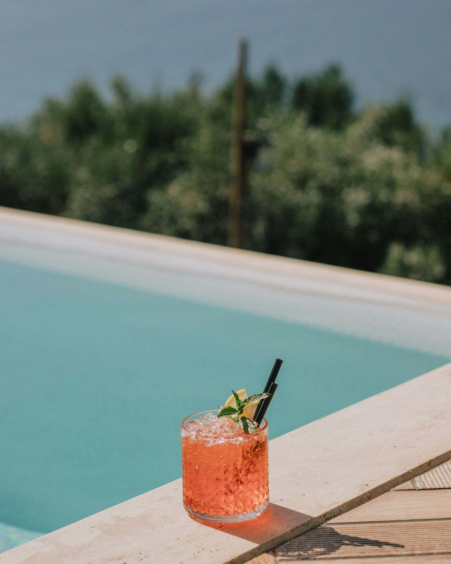 A refreshing coral-colored cocktail with crushed ice and garnish sits on the edge of a turquoise swimming pool.