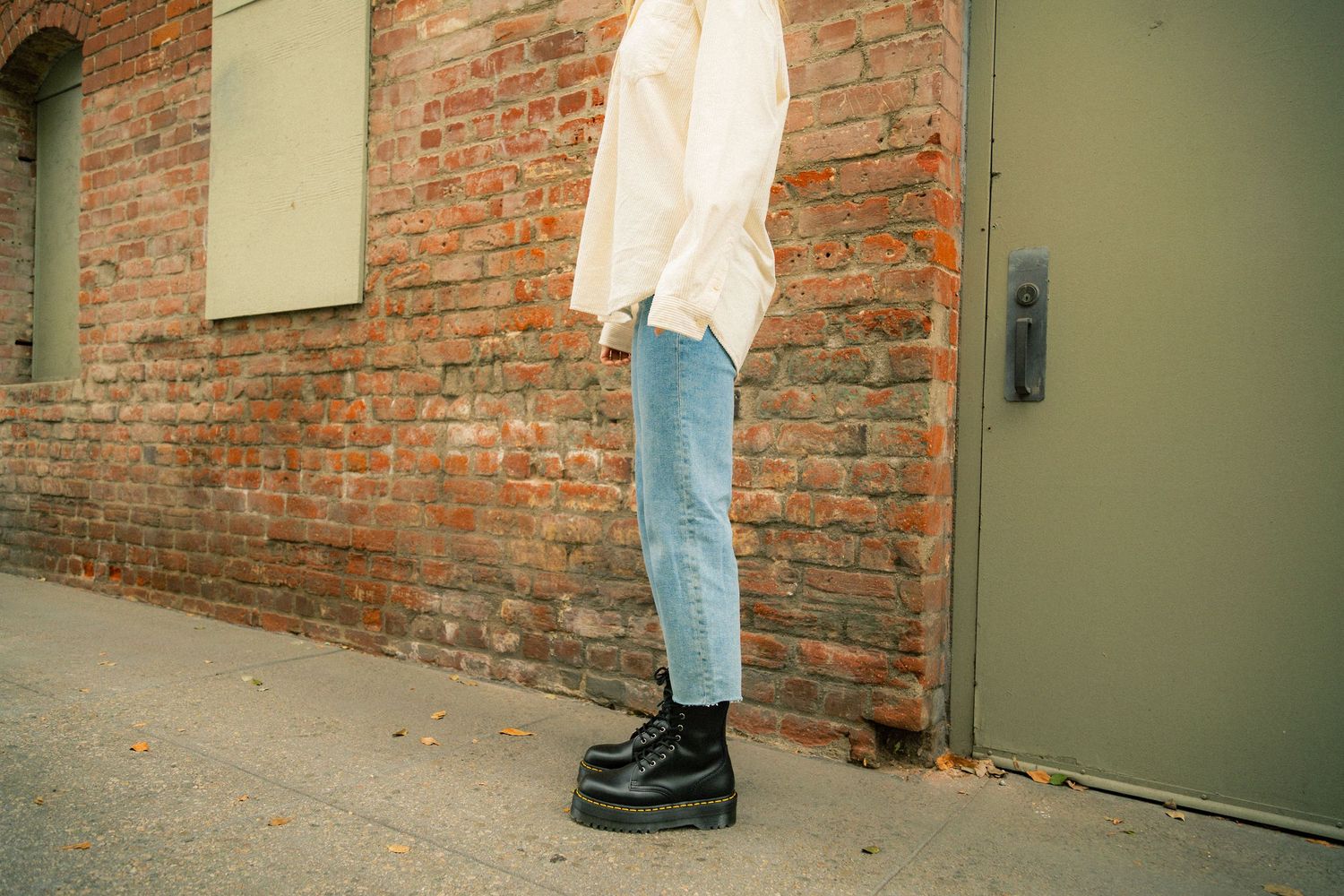 Person standing against a rustic red brick wall wearing light blue denim and dark shoes.