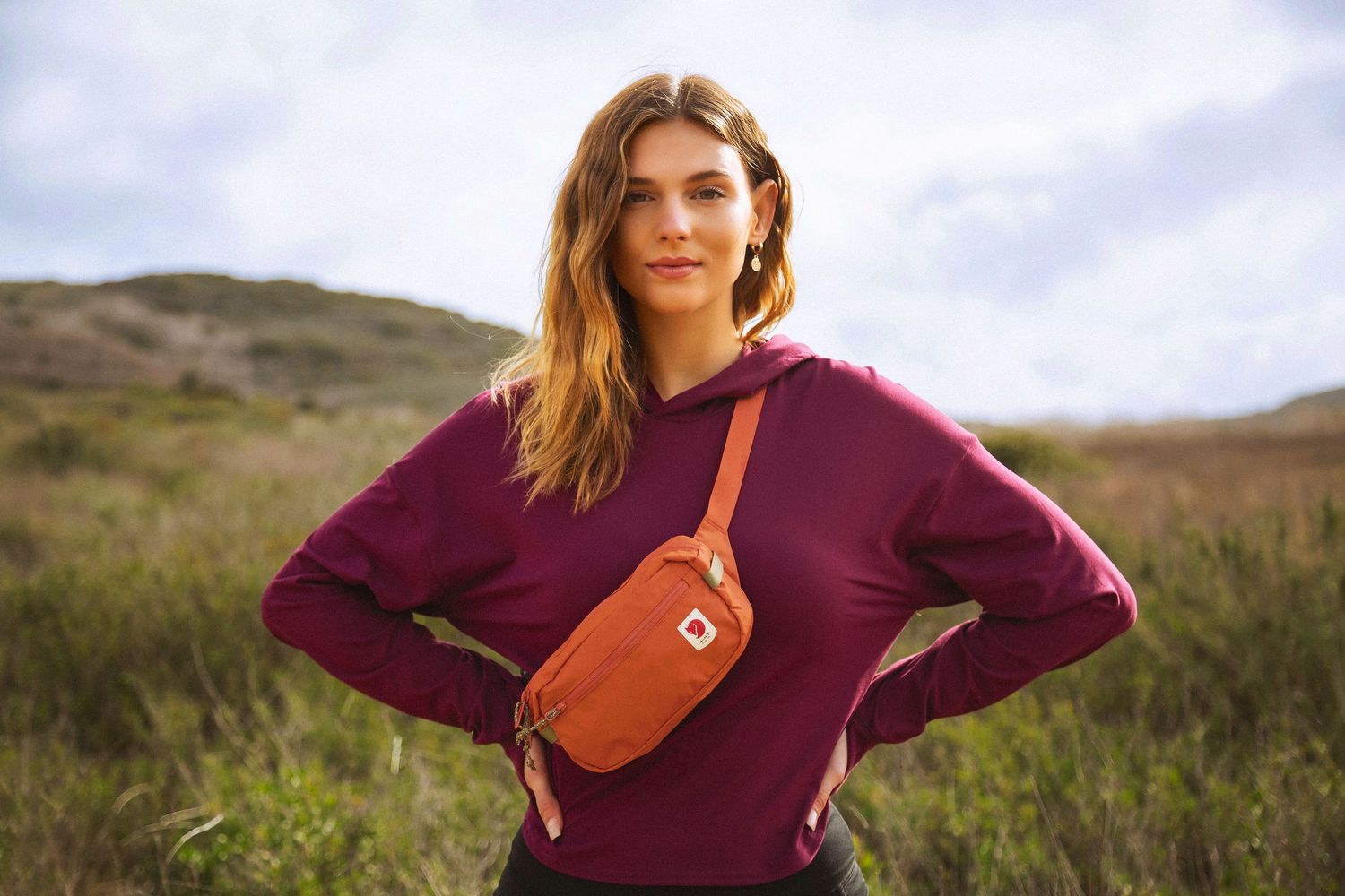 A woman in a burgundy sweater poses outdoors with a brown leather crossbody bag against a natural landscape.