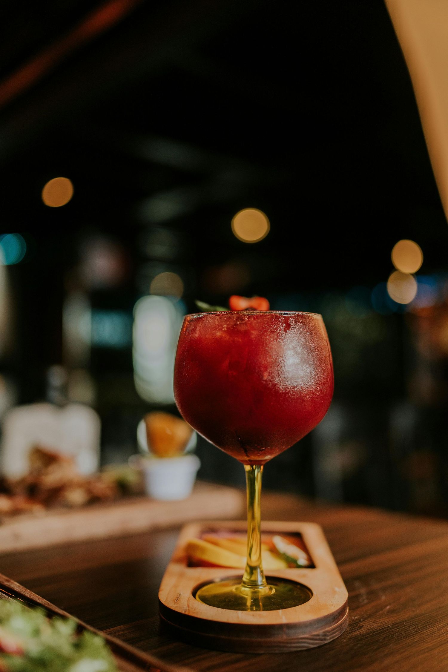 A red frozen cocktail served in a coupe glass sits on a dark wooden bar surface with blurred background lighting.