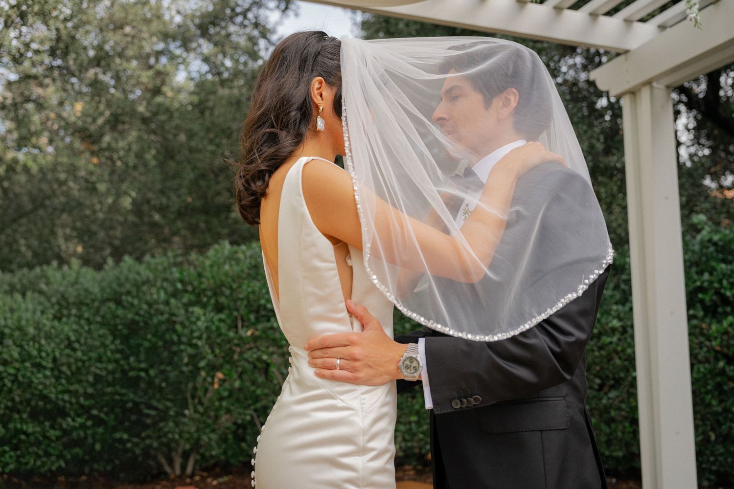 A romantic wedding couple embraces under a white pergola surrounded by lush greenery.